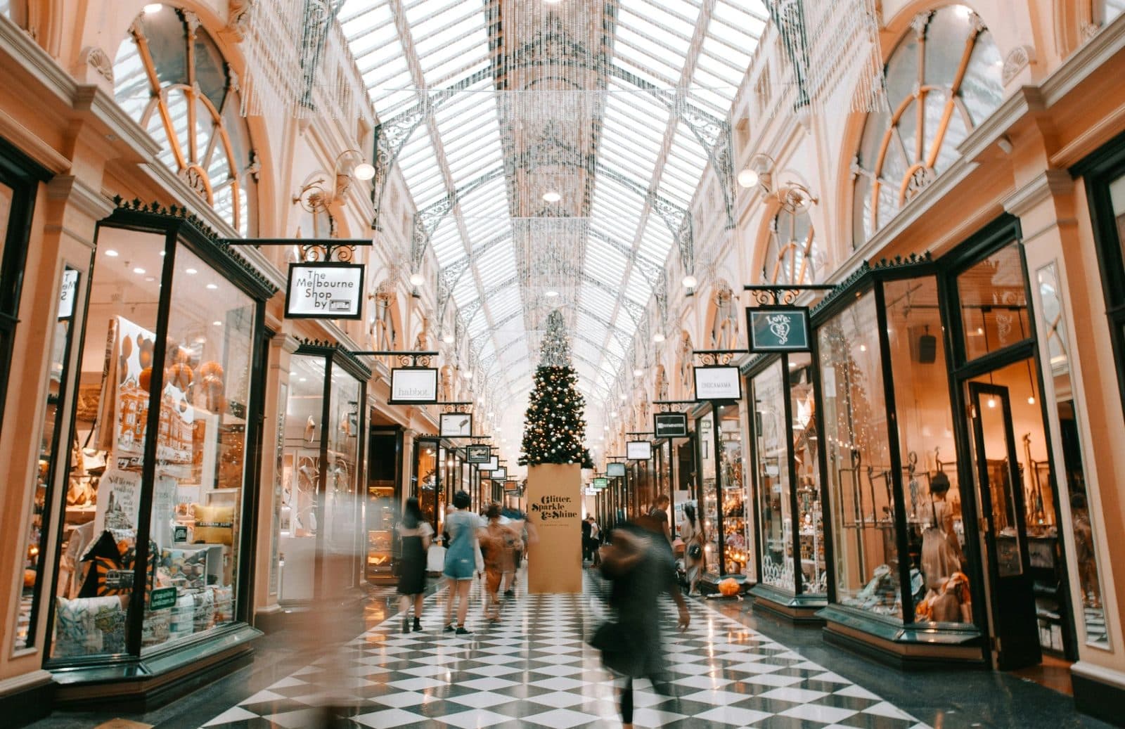 Shoppers walking through a Melbourne shopping arcade with a glass roof, a chequered tiled floor and small shopfronts on both sides